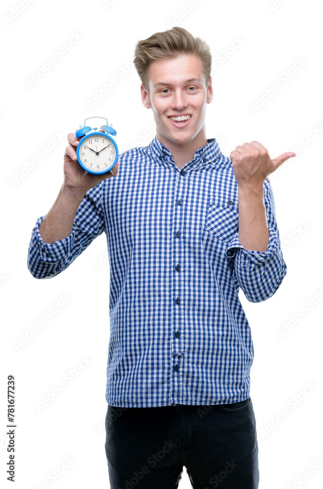 Wall mural Young handsome blond man holding alarm clock pointing with hand and finger up with happy face smiling