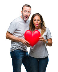 Middle age hispanic casual couple in love holding red heart over isolated background scared in shock with a surprise face, afraid and excited with fear expression