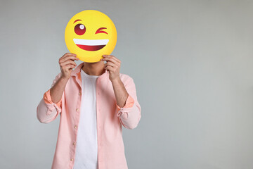 Man covering face with happy winking emoticon on grey background. Space for text