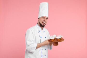 Happy professional confectioner in uniform holding delicious cupcakes on pink background