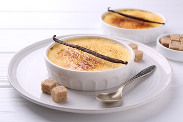 Delicious creme brulee in bowls, vanilla pods, sugar cubes and spoon on white wooden table, closeup