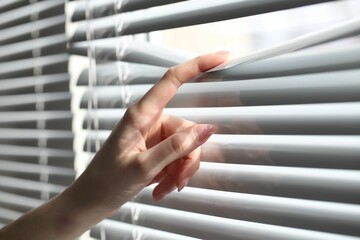 Woman separating slats of white blinds indoors, closeup