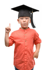 Dark haired little child wearing graduation cap surprised with an idea or question pointing finger with happy face, number one