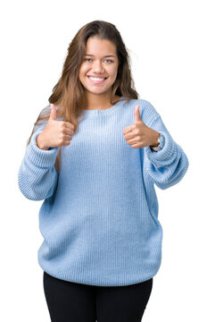 Young beautiful brunette woman wearing blue winter sweater over isolated background success sign doing positive gesture with hand, thumbs up smiling and happy. Looking at the camera.