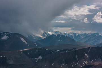 Dark atmospheric landscape with mountain silhouettes and large snow-capped peaked top in rainy low clouds. Dramatic alpine view to high mountains in gray cloudy sky. Big snowy mountain range in rain.