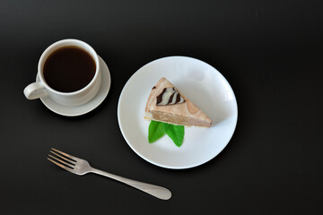 A piece of cream cheesecake with mint on a plate, a fork and a cup of hot black coffee on a saucer on a black background.