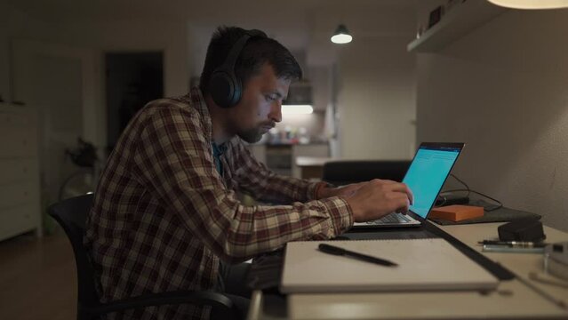 Focused man in headphones with a laptop late at night at home, taking notes, student learning a language, watching an online webinar, hearing an audiocourse, the concept of e-education. Homework. 