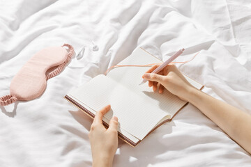 Woman writing on blank white page diary journal on white bedcloth in bed, holds paper notebook and pen in hands, device free time before sleep or morning, digital detox, psychological wellbeing