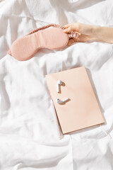 Woman hand takes pink eye sleep mask, near headphones, notebook or wish diary in bed, top view minimal lifestyle photo, copy space, Increasing mindfulness, well sleep, home relaxation concept