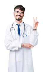 Fototapeta premium Young doctor man wearing hospital coat over isolated background smiling with happy face winking at the camera doing victory sign. Number two.