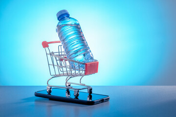 Plastic bottle in shopping trolley, smartphone on  dark grey desk. Application for ordering bottled purified water.