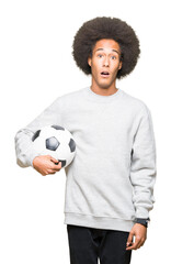 Young african american man with afro hair holding soccer football ball scared in shock with a...