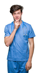 Young doctor wearing medical uniform over isolated background with hand on chin thinking about question, pensive expression. Smiling with thoughtful face. Doubt concept.