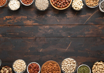 Mixed assorted nuts and seeds in various bowls on dark wooden table and space for text in the middle.Top view.