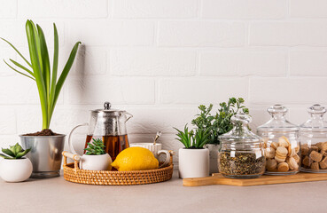 Stylish kitchen background with glass tea pot, lemon, cup, tea jars, sugar, meringue pastry. Green plants in pots. Front view. White brick wall.