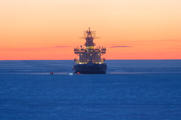 Icebreaker in the frozen sea. Beautiful winter arctic landscape. Icebreaker among the ice-covered sea expanses. Cars on the ice near a ship anchored in the roadstead. Evening twilight after sunset. - 781818071