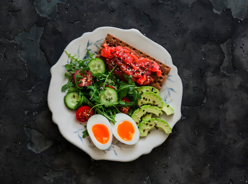 Delicious breakfast - salmon rye cracker sandwich, boiled egg, salad, avocado on a dark background, top view