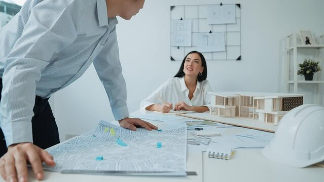 Skilled caucasian interior designer working with house model while discussing with asian architect engineer about house plan on meeting table with blueprint and house model scatter around. Immaculate.