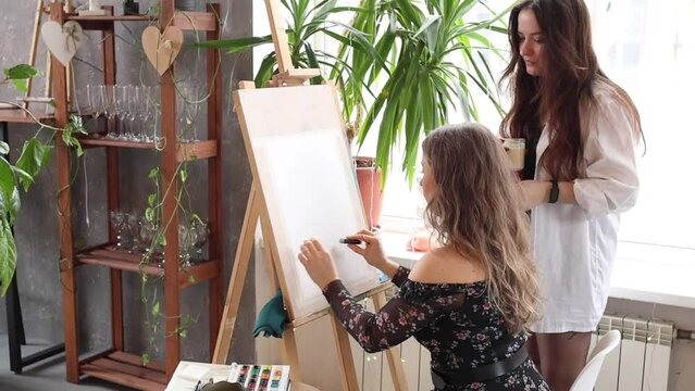 Two women working in art studio. Female artist girls standing in front of wooden easel. Beautiful teacher and student painting on paper in class. Creative process