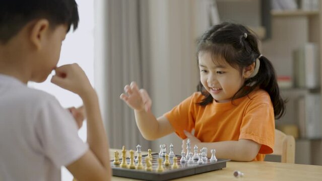 Two children playing a game of chess. One is smiling and the other is frowning. The boy in the white shirt is smiling and the girl in the orange shirt is frowning