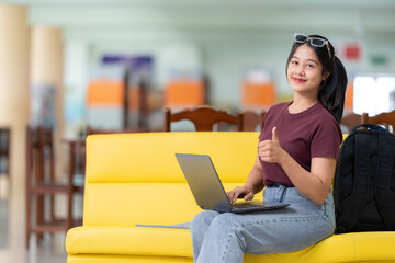 Asian female student with long hair studying online with a notebook computer wearing glasses and casual clothes Thumbs up, Like symbol in the school library, carrying a bag, sitting on a yellow sofa