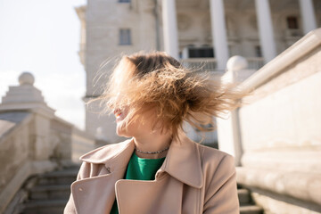 A woman with long hair is standing on a set of stairs. She is wearing a green shirt and a tan coat.