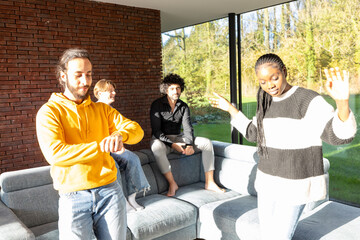 In a living room bathed in sunlight, a diverse group of friends is engaged in a lively conversation. One man checks the time, perhaps indicating it's time for an event, while a woman gestures