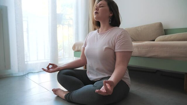 Woman meditating in lotus position with eyes closed, serene home environment.