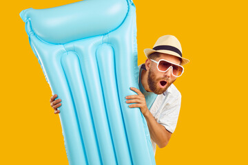 Portrait of excited shocked funny young man in sunglasses and beach hat holding inflatable mattress with open mouth on studio yellow background. Summer holiday trip and travel concept. - obrazy, fototapety, plakaty