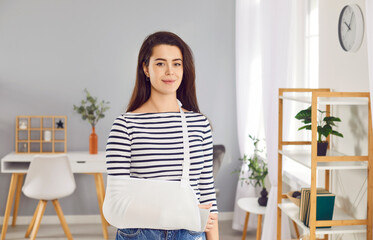  Smiling woman stands with bandage on her arm, showing strength and endurance despite injury. Portrait of smiling woman in stylish home interior wearing medical brace to support broken arm.