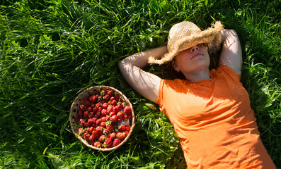 Happy  woman wearing a straw hat covering his face and an orange T-shirt laying on grass with a strawberry basket in summer. 