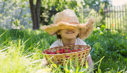 Happy  adorable little boy wearing a straw hat covering his face laying on grass with a strawberry basket in summer. Happy little boy having fun outdoors.