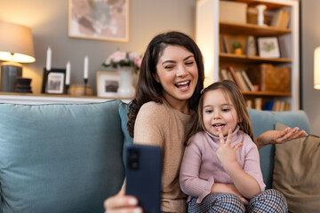 Smiling mother and daughter on video call using digital phone at home. while making video call and waving hello sitting on sofa. Woman and her cute little kid doing video conference with grandparents.