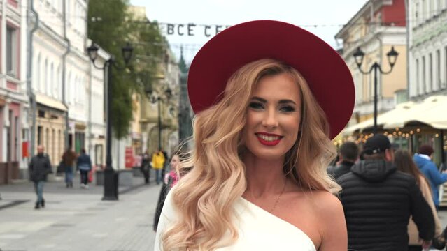  stunning blonde woman in a beautiful white dress is seen walking down a city street. She's making a strong fashion statement with a bold red hat and matching red heels. she turns around. 