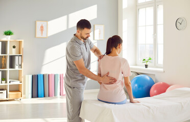 Male osteopath examining young woman in modern office. Young physiotherapist doing healing treatment on woman back in rehabilitation clinic. Chiropractor helping patient with scoliosis and back pain. - obrazy, fototapety, plakaty