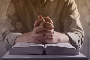 Religion. Christian man praying over Bible at table, closeup. Bokeh effect