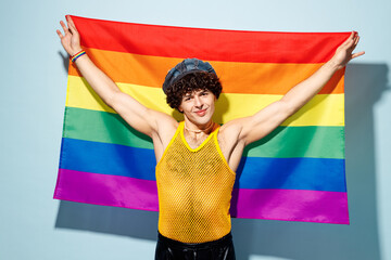 Young happy gay Latin man wear mesh tank top hat clothes raise up hands with striped rainbow flag...