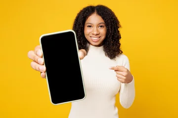 Poster Im Rahmen Little fun kid teen girl of African American ethnicity wear white casual clothes hold use point on blank screen area mobile cell phone isolated on plain yellow background. Childhood lifestyle concept. © ViDi Studio