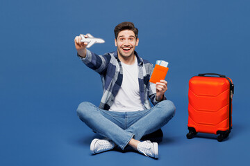 Traveler man wears casual clothes sit near bag hold passport ticket air plane mockup isolated on plain blue background. Tourist travel abroad in free time rest getaway Air flight trip journey concept