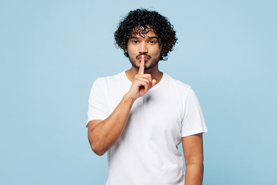 Young secret Indian man he wear white t-shirt casual clothes say hush be quiet with finger on lips shhh gesture isolated on plain pastel light blue cyan background studio portrait. Lifestyle concept.