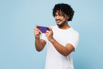 Young gambling Indian man wear white t-shirt casual clothes use play racing app on mobile cell phone hold gadget smartphone for pc video games isolated on plain blue cyan background Lifestyle concept