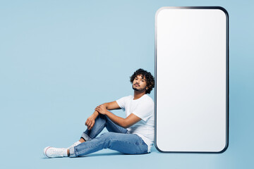 Full body sideways young Indian man wear white t-shirt casual clothes sits near big huge blank screen mobile cell phone smartphone with area isolated on plain pastel blue background Lifestyle concept