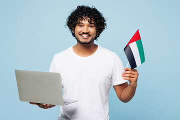 Young happy IT Indian man he wear white t-shirt casual clothes hold flag of UAE use work on laptop...