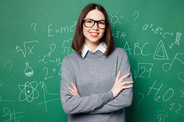 Young happy smart teacher woman wear grey casual shirt glasses hold hands crossed folded look camera isolated on green wall chalk blackboard background studio Education in high school college concept