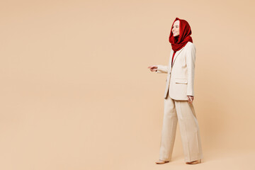 Full body side view smiling happy young Arabian Asian Muslim woman wearing red abaya hijab suit clothes walk go look aside isolated on plain beige background studio. UAE middle eastern Islam concept. - obrazy, fototapety, plakaty