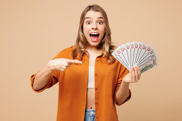 Young happy woman she wears orange shirt casual clothes hold point finger on fan of cash money in dollar banknotes do winner gesture isolated on plain pastel light beige background. Lifestyle concept.