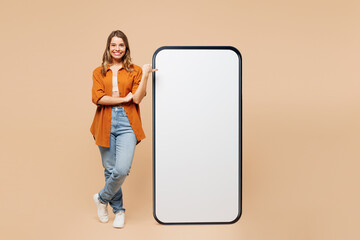 Full body young woman she wears orange shirt casual clothes point thumb finger on big huge blank screen mobile cell phone smartphone with mockup area isolated on plain pastel light beige background.
