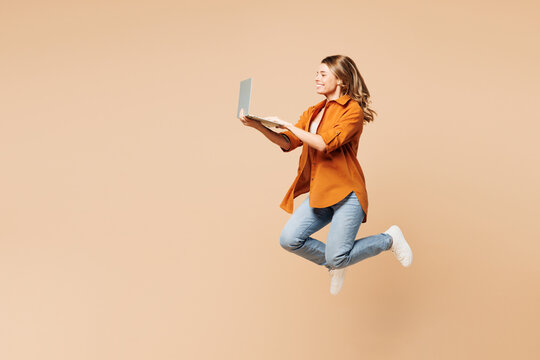 Full body side view young IT woman she wear orange shirt casual clothes jump high hold use work on laptop pc computer isolated on plain pastel light beige background studio portrait Lifestyle concept