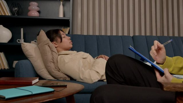 Stressed charming African American woman lying on sofa, talking with caring female mental health professional about fears and certain mental disorders during hypnotherapy session at doctor office.