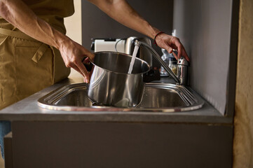Close-up of a man in beige chef's apron, standing at the kitchen sink and filling a pot of water to...